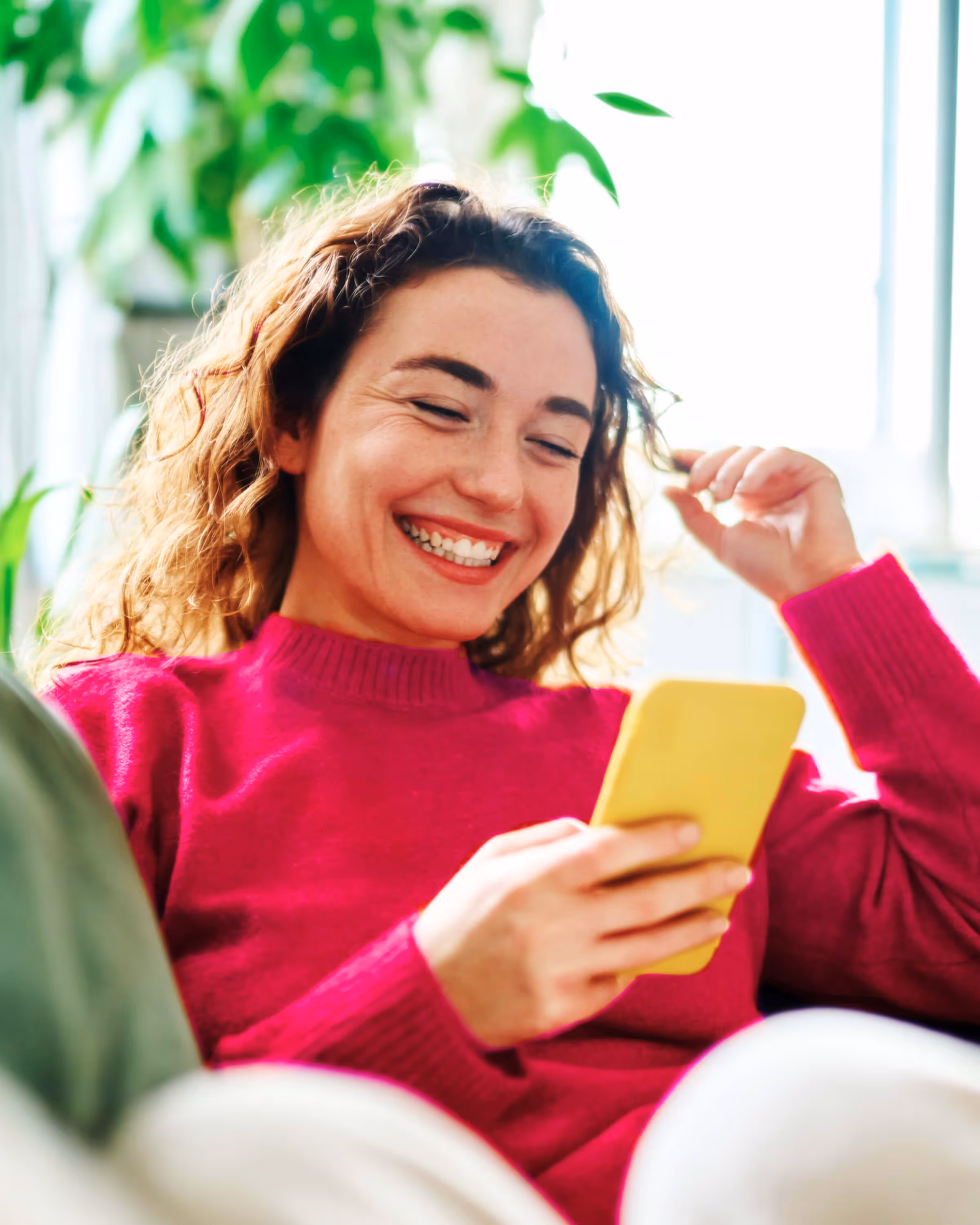 Smiling woman in a bright pink sweater looking at her yellow smartphone while sitting indoors with green plants in the background.