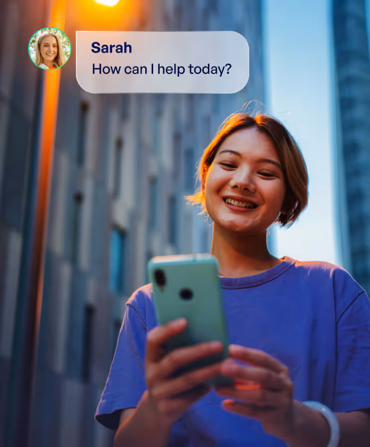 Smiling young woman in a purple shirt looking at her phone outdoors at dusk, with a message bubble saying 'Sarah How can I help today?'
