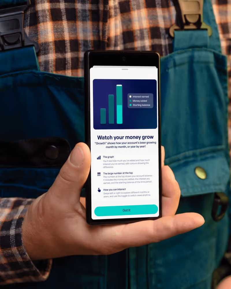 Person in plaid shirt and green overalls holding a smartphone displaying a financial app screen with a bar chart showing interest earned, money added, and starting balance, titled 'Watch your money grow'.