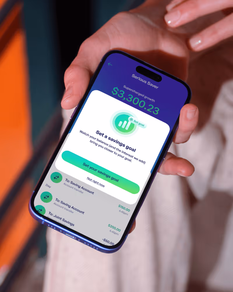 Hand holding a smartphone displaying a savings app with a $3,300.23 balance and an option to set a savings goal.