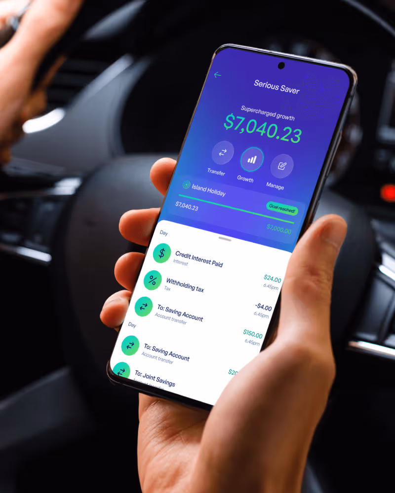 Smartphone showing a savings app with $7,040.23 balance labeled Serious Saver and an Island Holiday savings goal reached.
