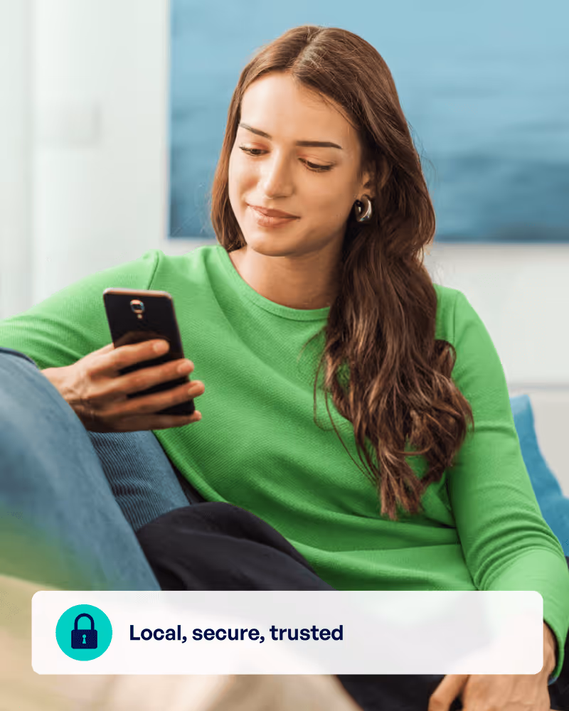 Young woman in a green sweater sitting on a couch and looking at her smartphone with a relaxed smile.