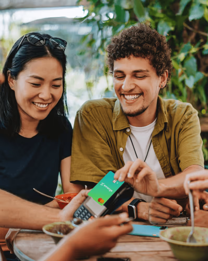 Smiling man making contactless payment with green Emergo card on card reader while sitting at wooden table with friends outdoors.