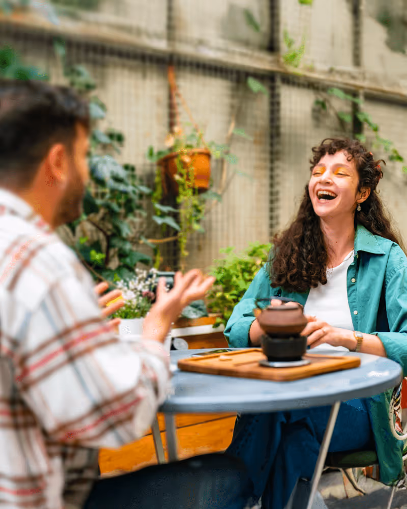 Two people sitting at a round table outdoors, smiling and chatting amid green plants, with a teapot on the table.