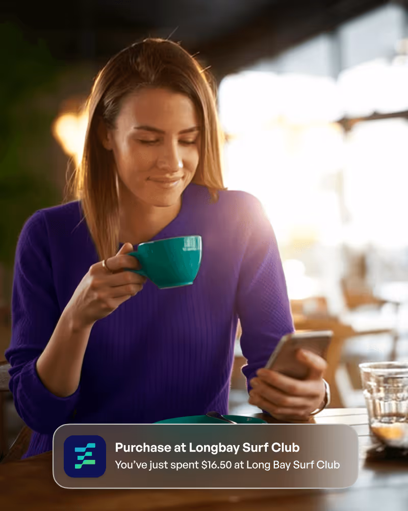A woman in a purple sweater drinking from a green cup while looking at her phone in a bright café, with a notification about a $16.50 purchase at Long Bay Surf Club.