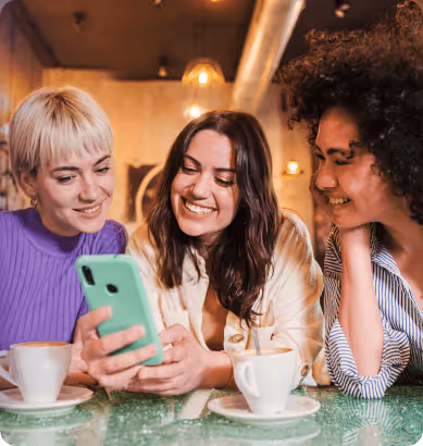 Three women smiling and looking at a smartphone while sitting at a table with coffee cups.