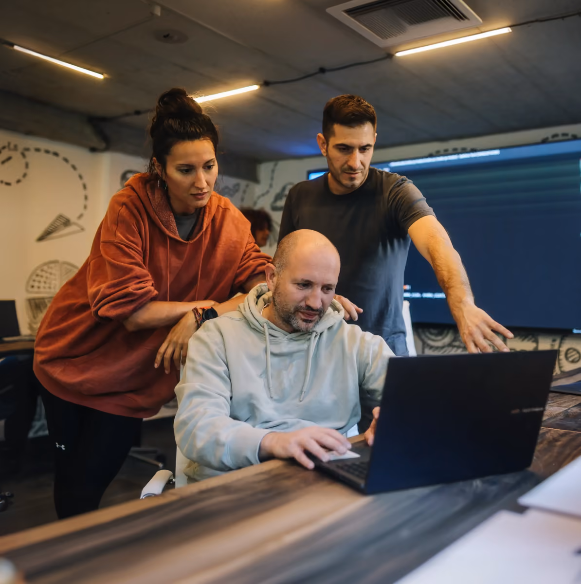 Three coworkers collaborate over a laptop in a modern office with doodles on the wall.