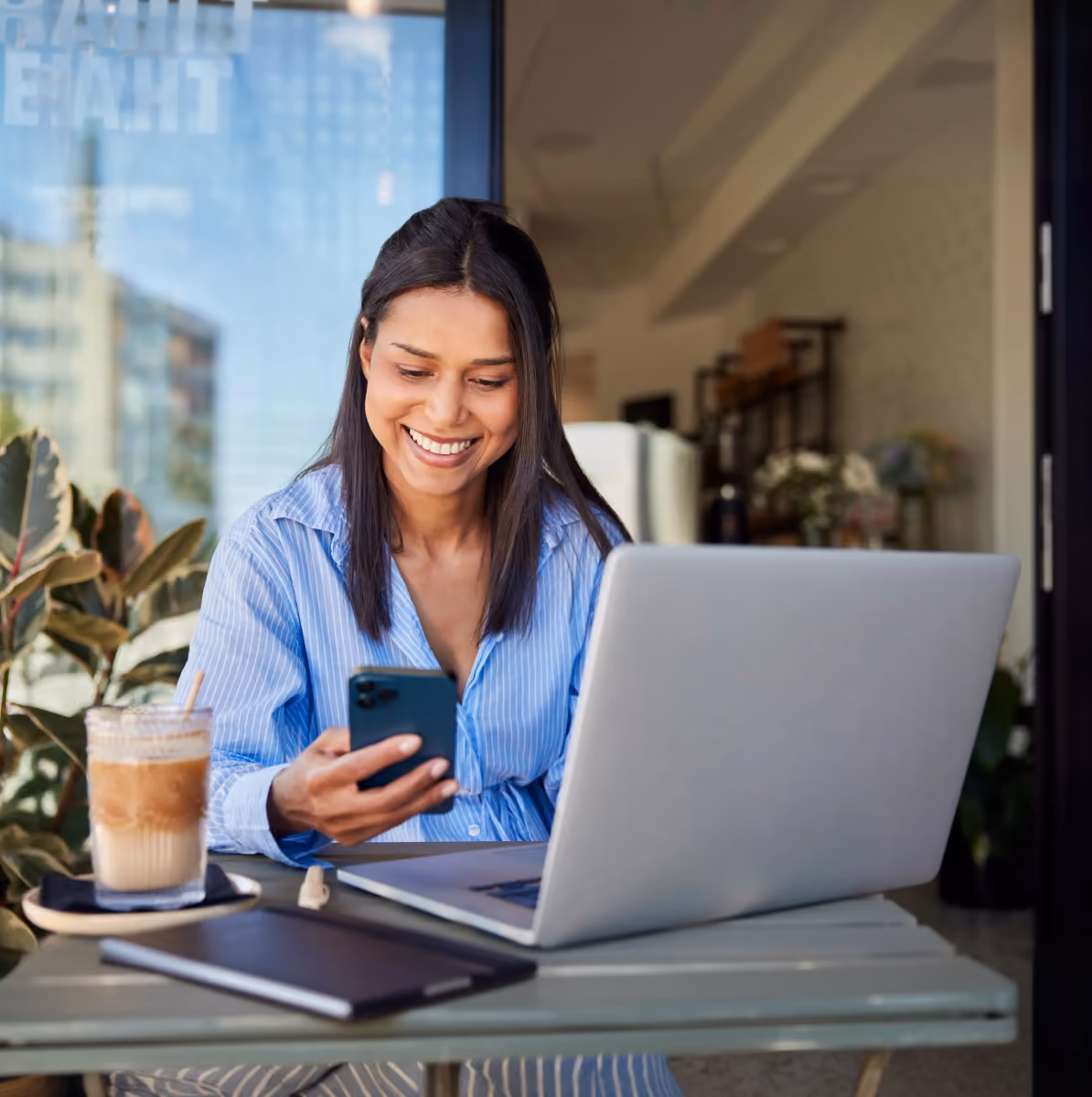 Smiling woman in blue shirt using smartphone and laptop at outdoor café table with iced coffee.