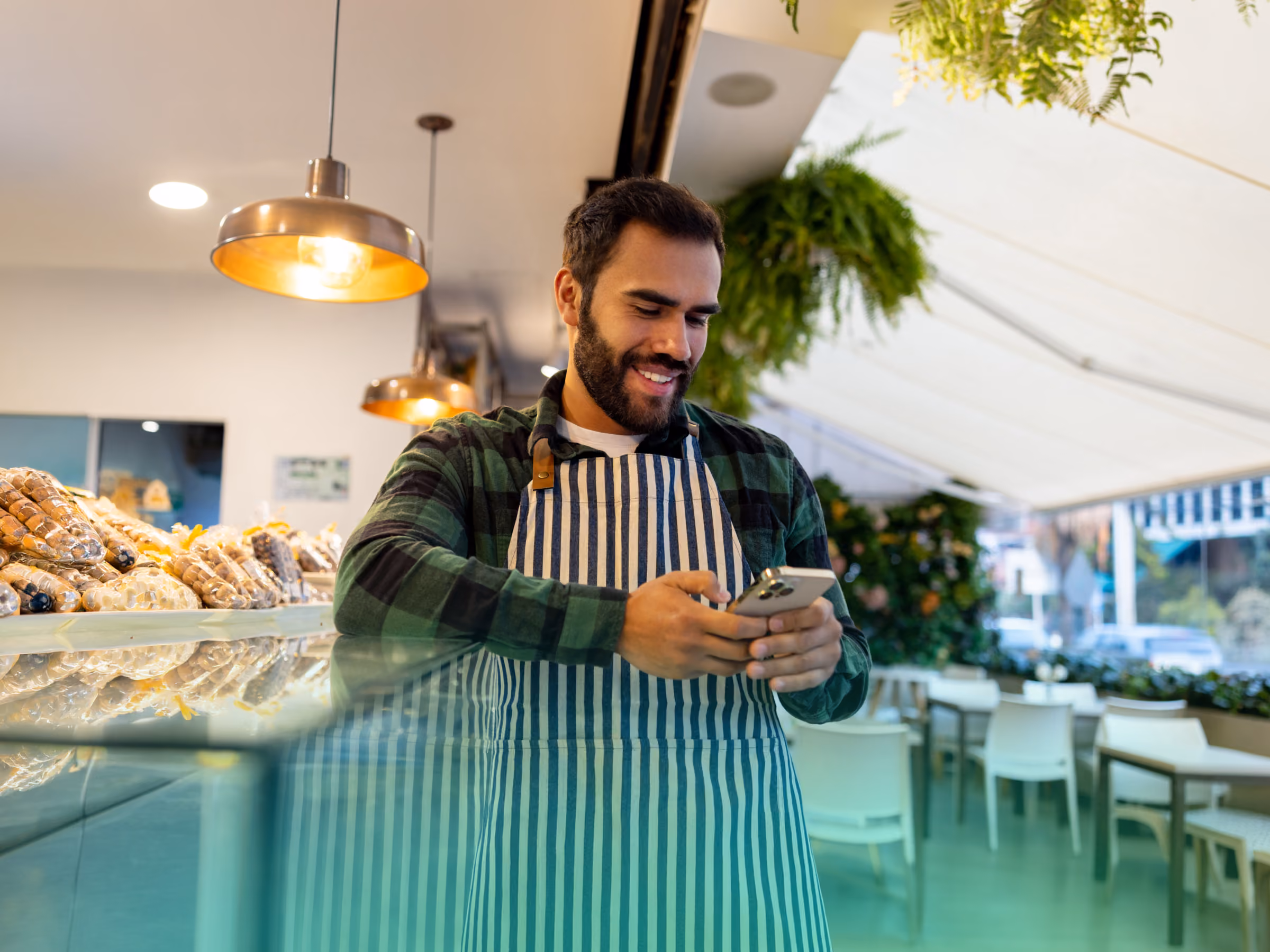 Smiling man in striped apron using smartphone behind bakery counter with pastries and plants.