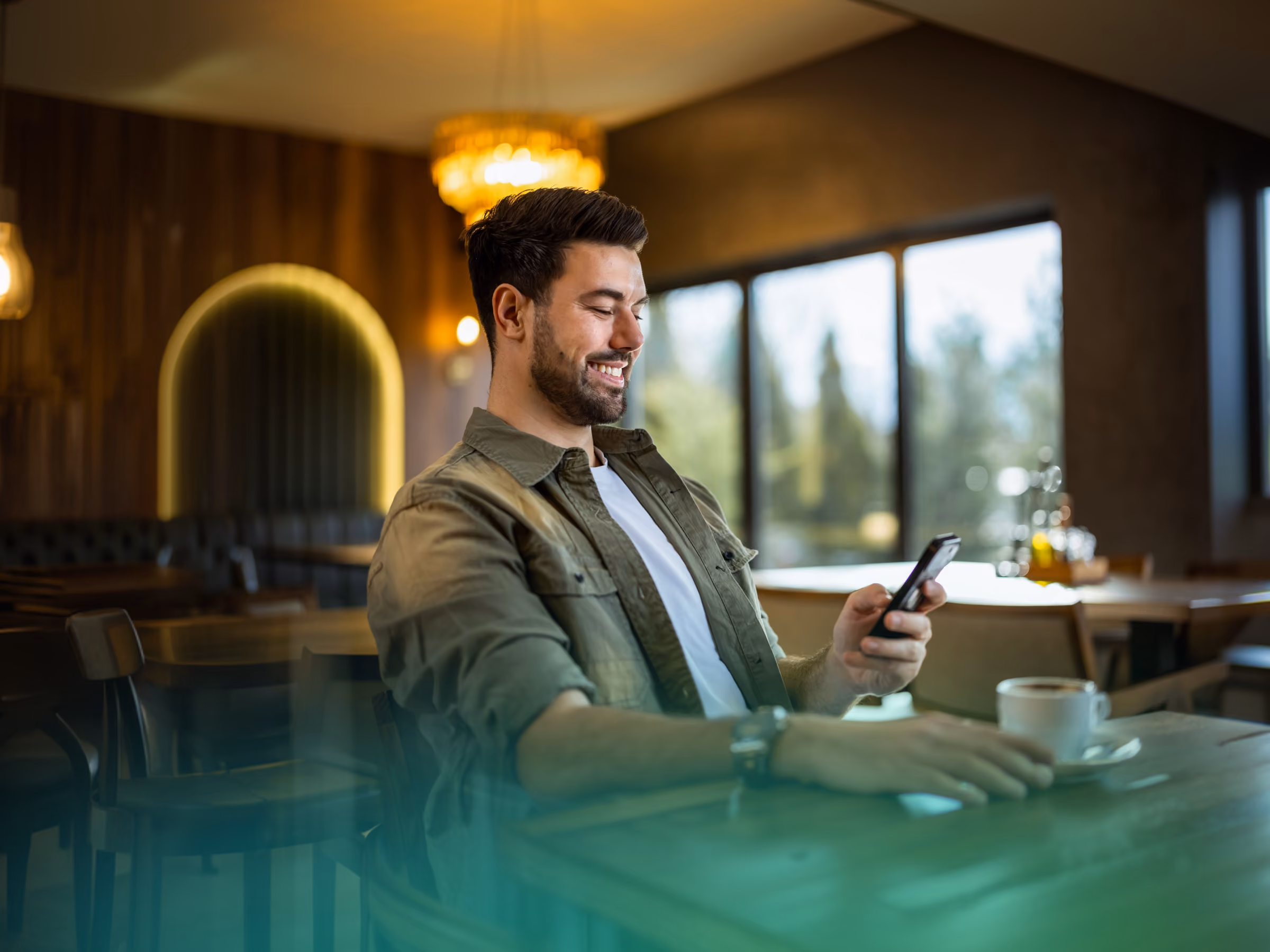 Smiling man with beard sitting at a table in a café, looking at his phone with a cup of coffee nearby.