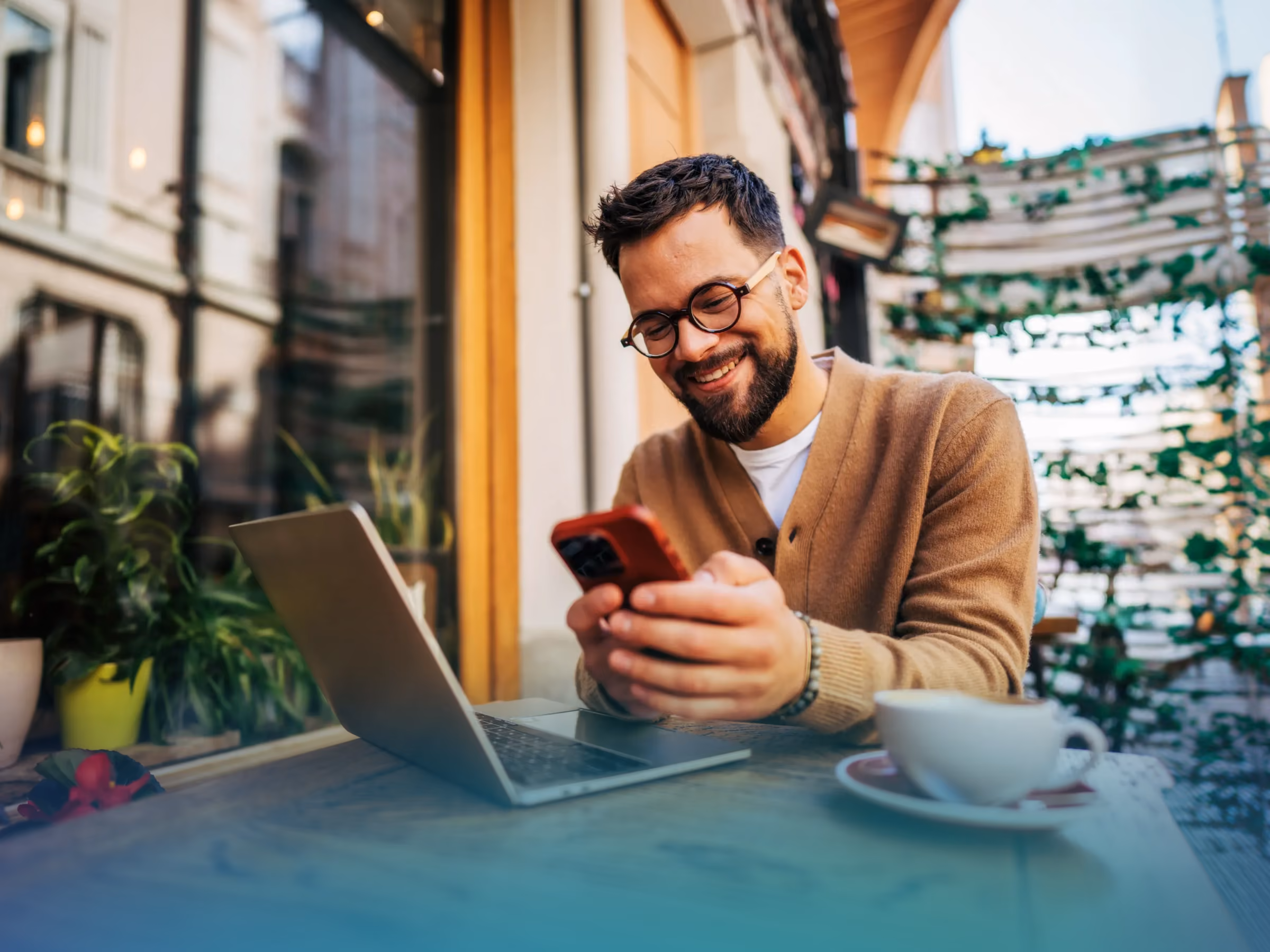 Smiling man with glasses using smartphone at outdoor café table with laptop and coffee cup.
