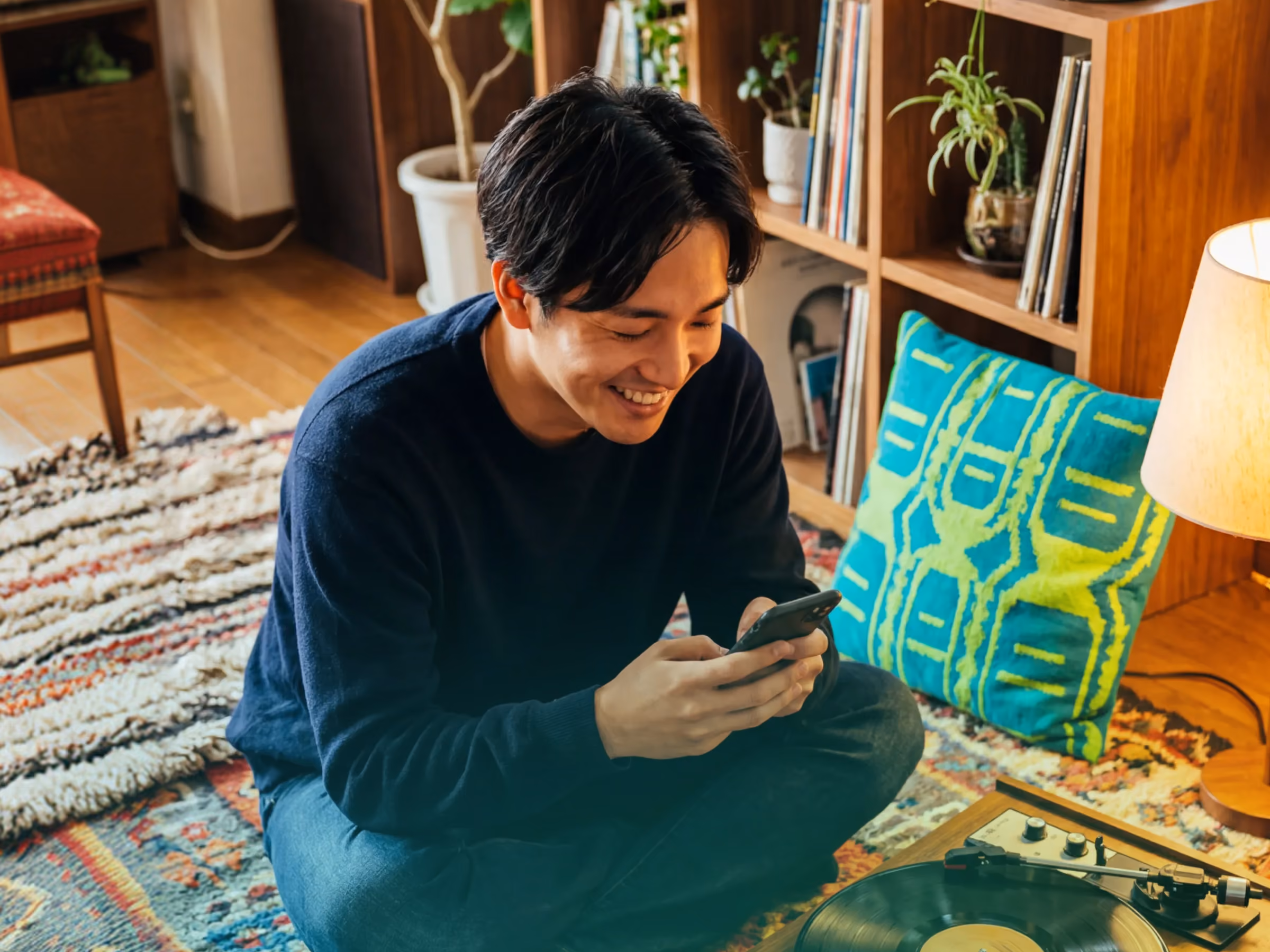 Smiling man sitting cross-legged on a rug, holding a phone near a turntable in a cozy room.