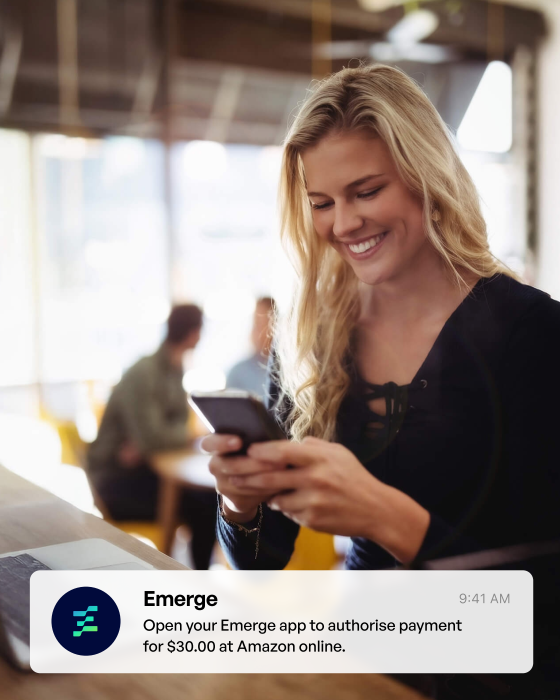 Smiling woman using her phone in a cafe, with an Emerge notification asking her to authorise a $30.00 payment at Amazon online.