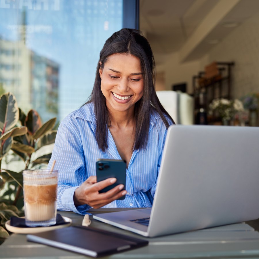 Smiling woman using her phone beside a laptop in a bright office.