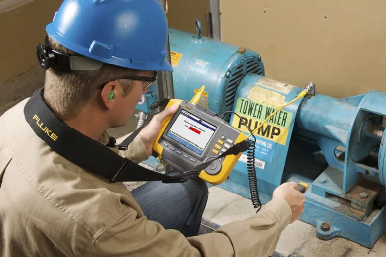 Technician wearing a blue hard hat using a handheld diagnostic device to test equipment near an industrial water pump.
