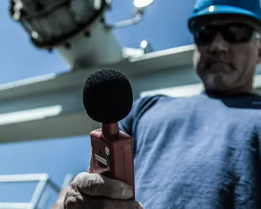 Industrial technician in safety gear holding a sound level meter during an acoustic measurement in an outdoor facility