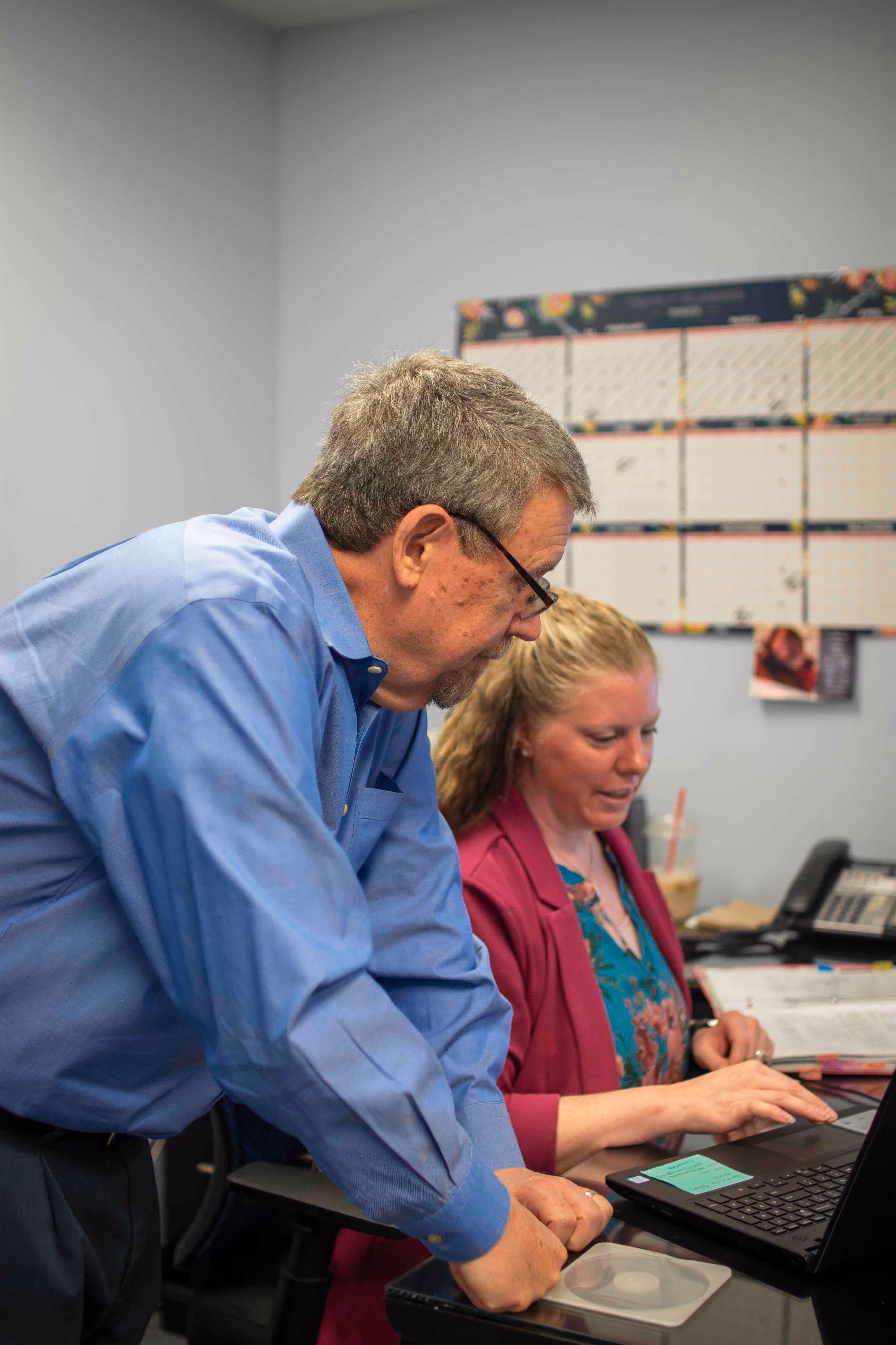 Man in blue shirt leaning over desk and woman in pink blazer using a laptop in an office.