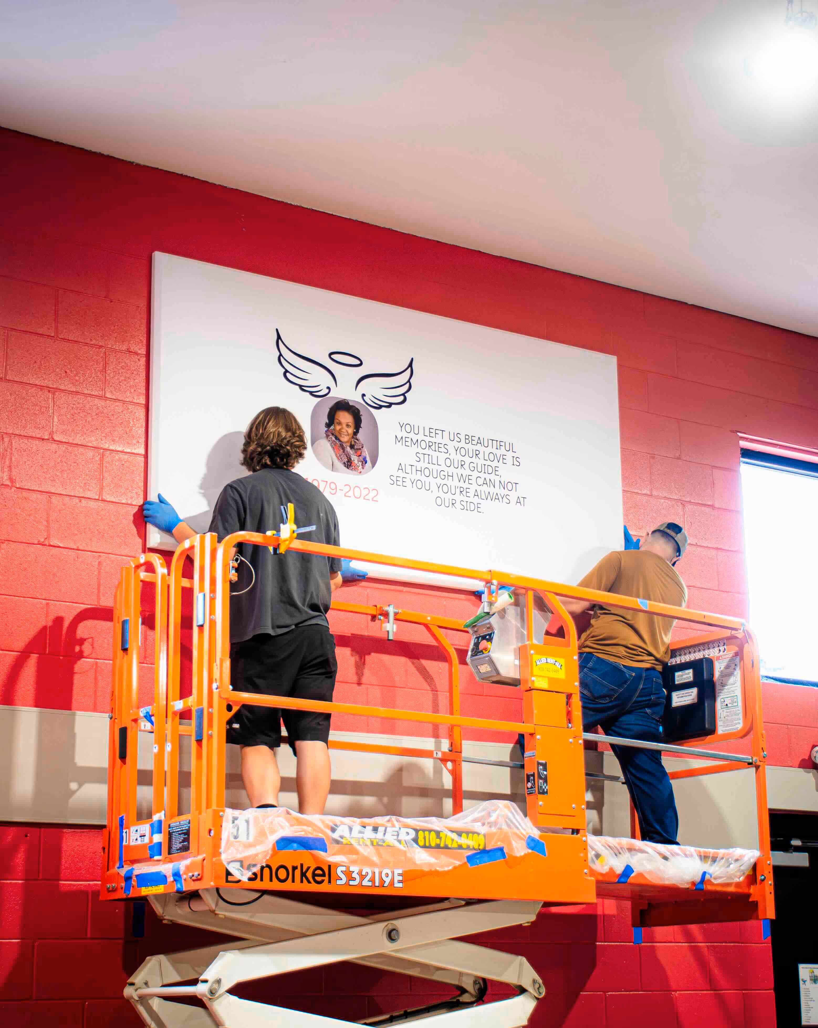 Two installers on a scissor lift mounting a memorial acoustic panel featuring a tribute photo and message on a red brick wall