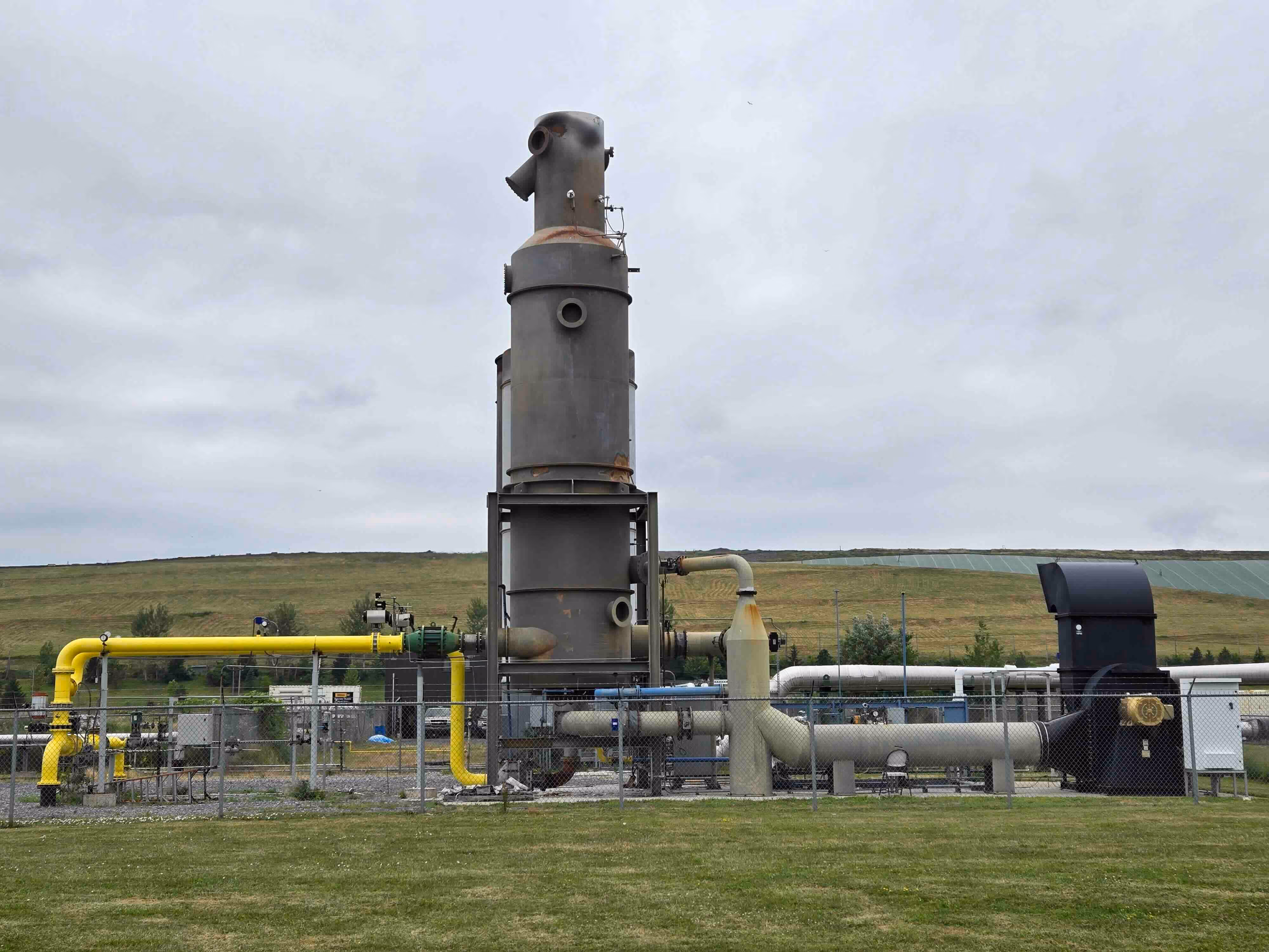 "Industrial gas processing equipment with vertical scrubber unit and yellow piping, enclosed in a fenced area at a landfill facility