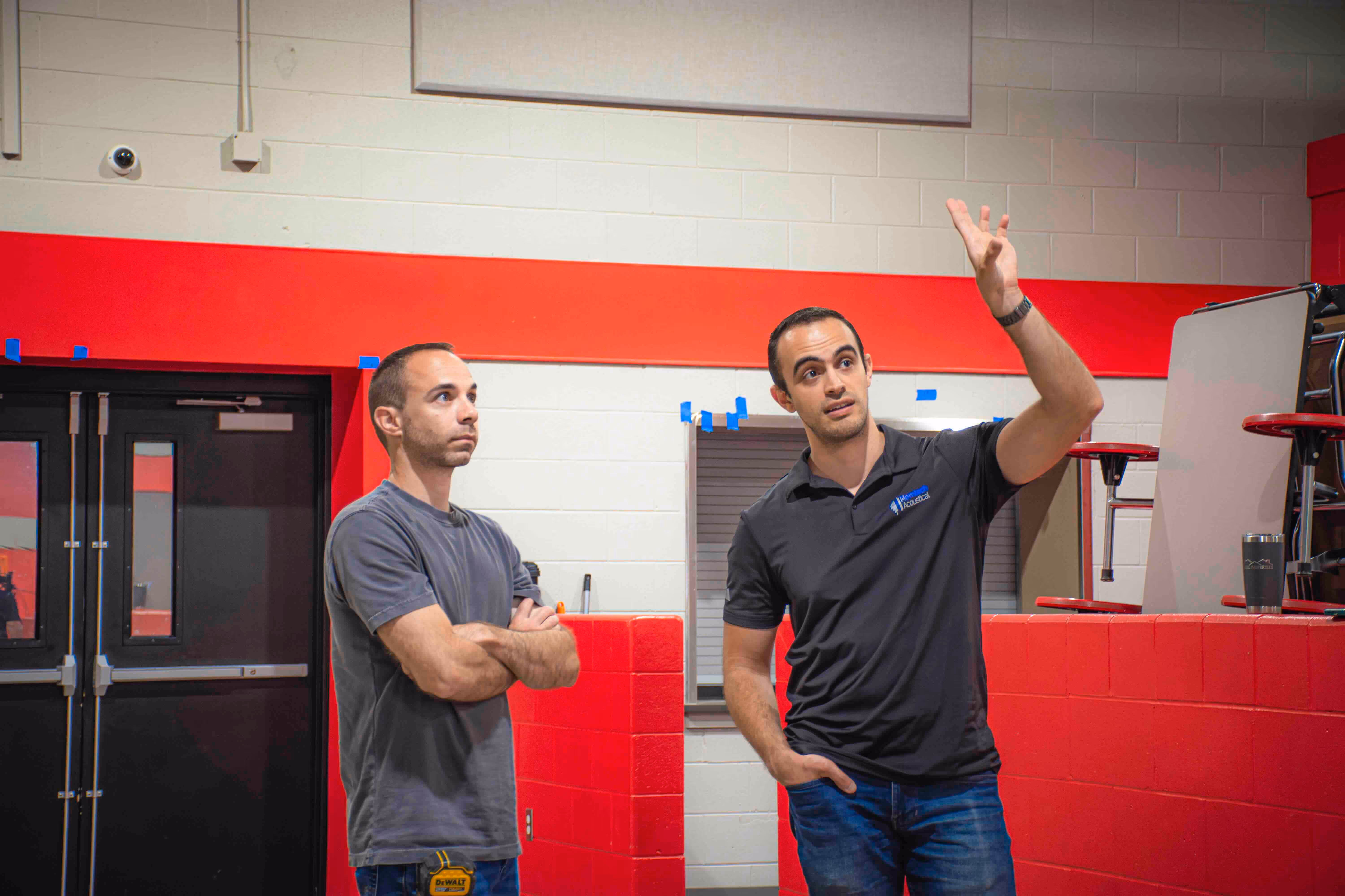 Two men discussing acoustic treatment plans inside a gymnasium, with one wearing a Memtech Acoustical shirt and pointing toward the wall panels