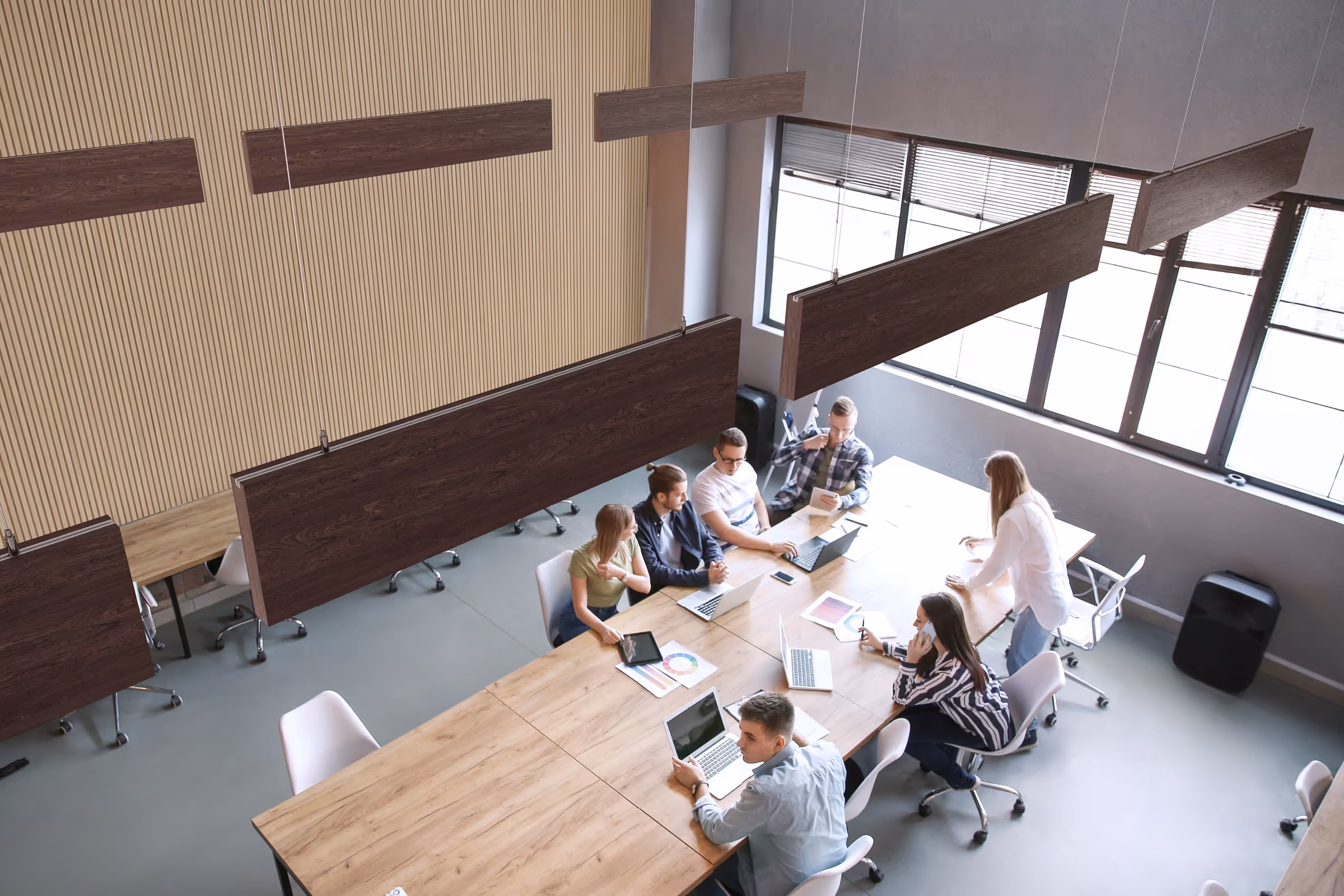 Modern office space with wood-finish acoustic baffles suspended from the ceiling, where a group of professionals are collaborating around a large conference table