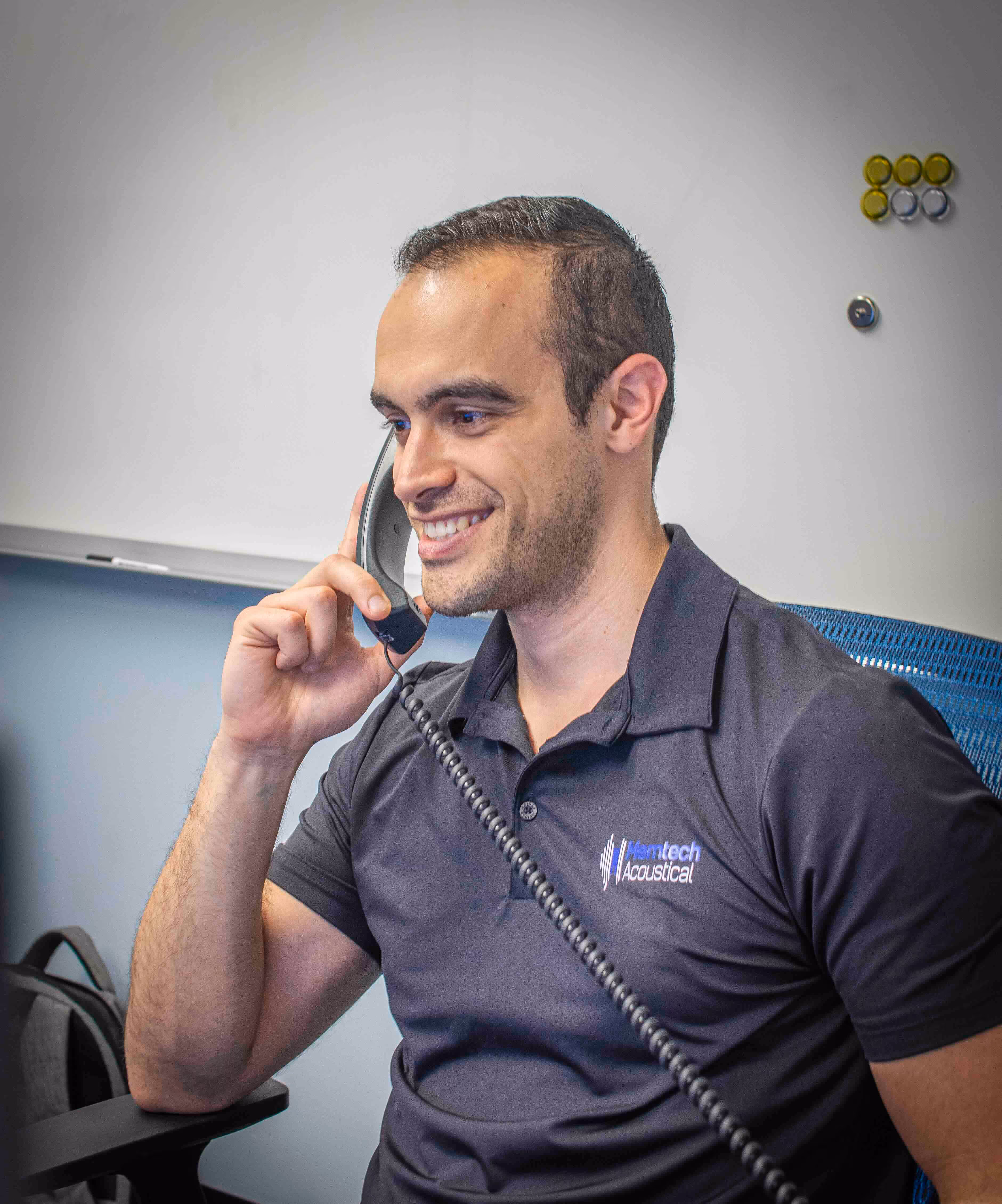 Smiling Memtech Acoustical team member, Michael Castano,  speaking on a landline phone in an office setting, wearing a branded polo shirt