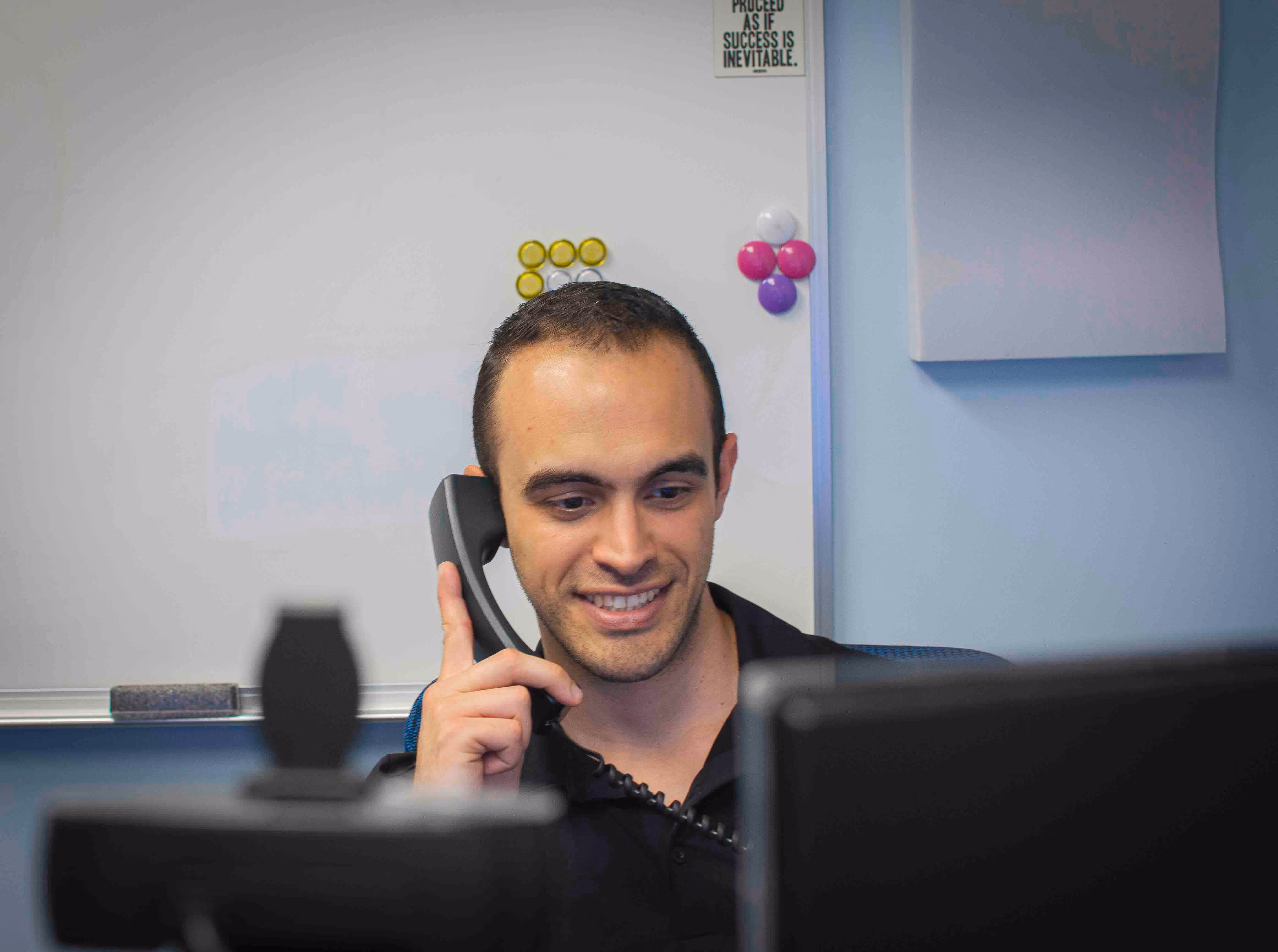 Memtech Acoustical team member smiling while speaking on the phone at his desk, with a whiteboard and motivational quote in the background