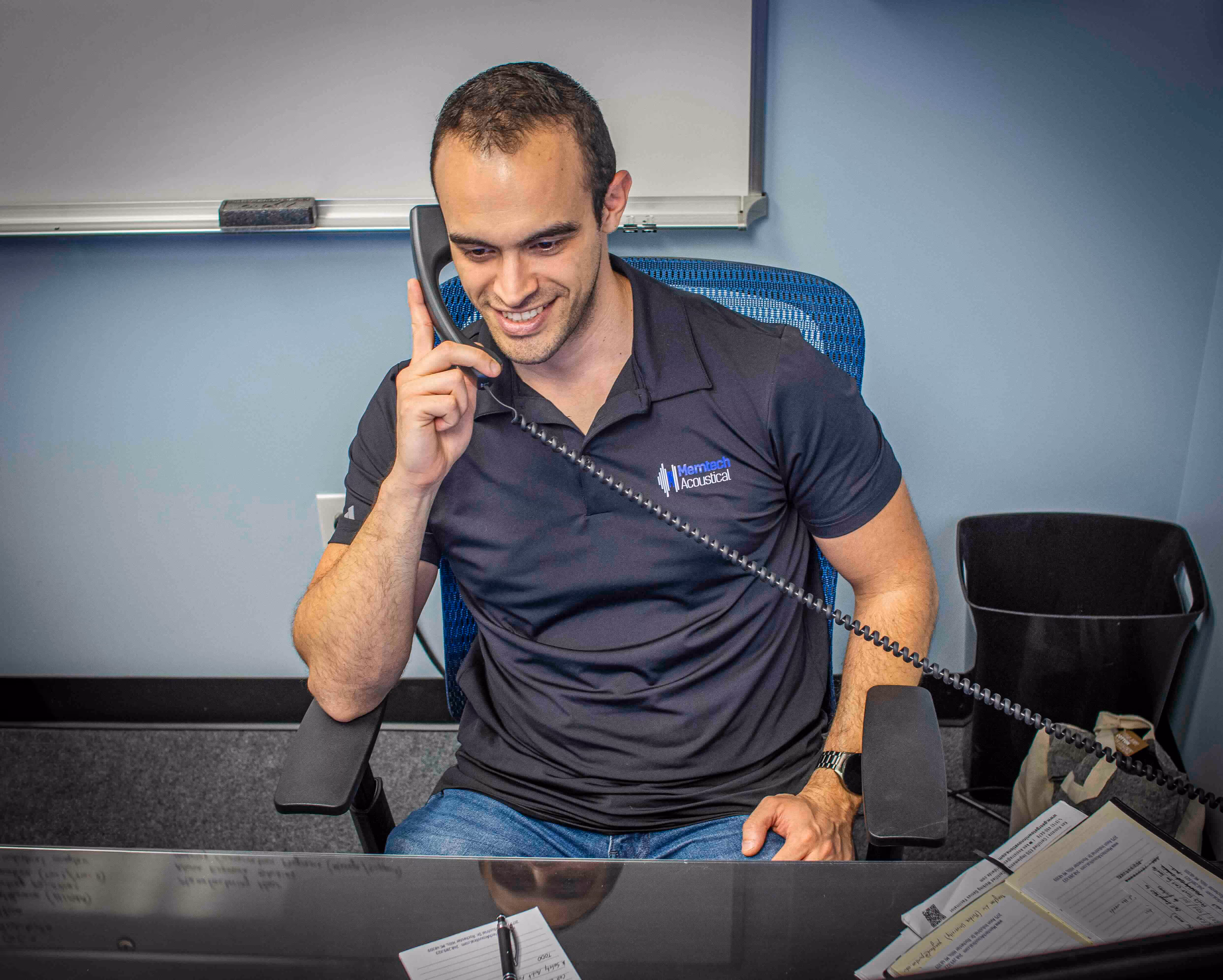 Office worker speaking on a corded phone while seated at a desk, wearing a branded polo shirt and smiling during the call.