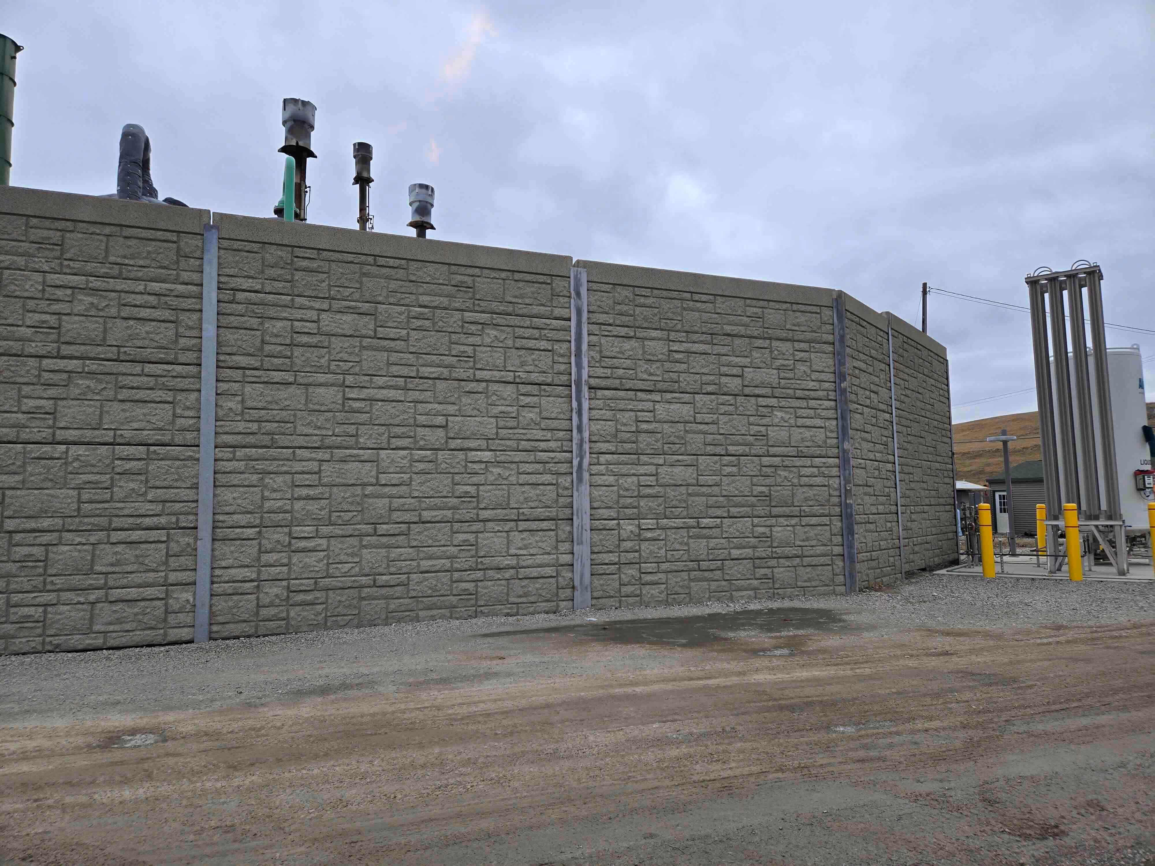 Tall sound barrier wall with textured concrete panels surrounding industrial equipment at a facility, designed for noise mitigation