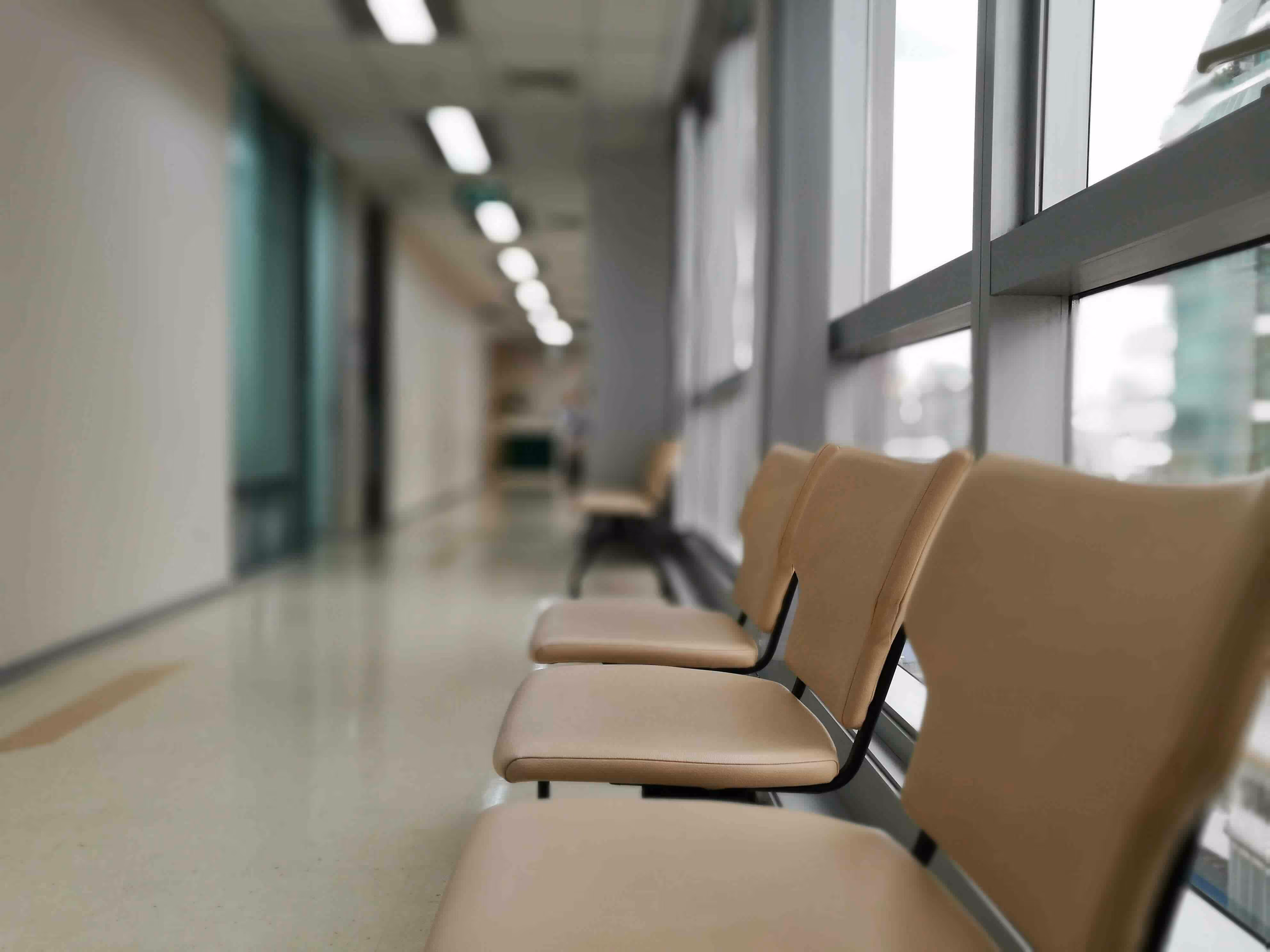 Empty chairs lined up in a brightly lit hospital or clinic hallway, with large windows providing natural light