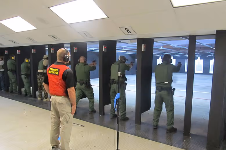 Group of law enforcement officers training at an indoor shooting range while an instructor in a red vest observes.