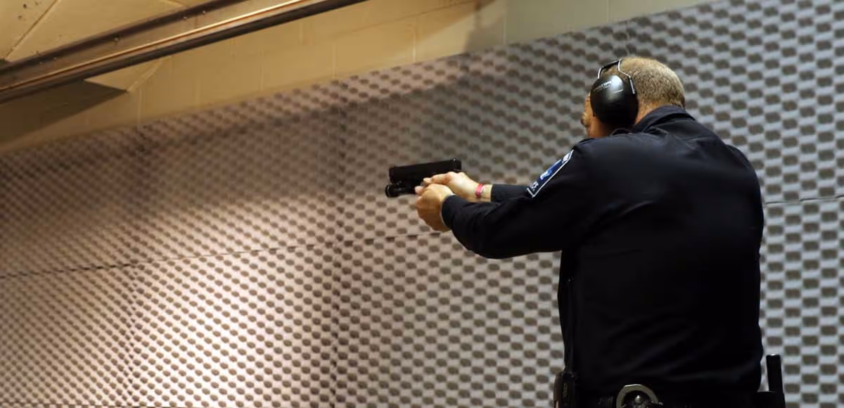 Police officer wearing ear protection aiming a handgun at an indoor shooting range with acoustic wall panels.