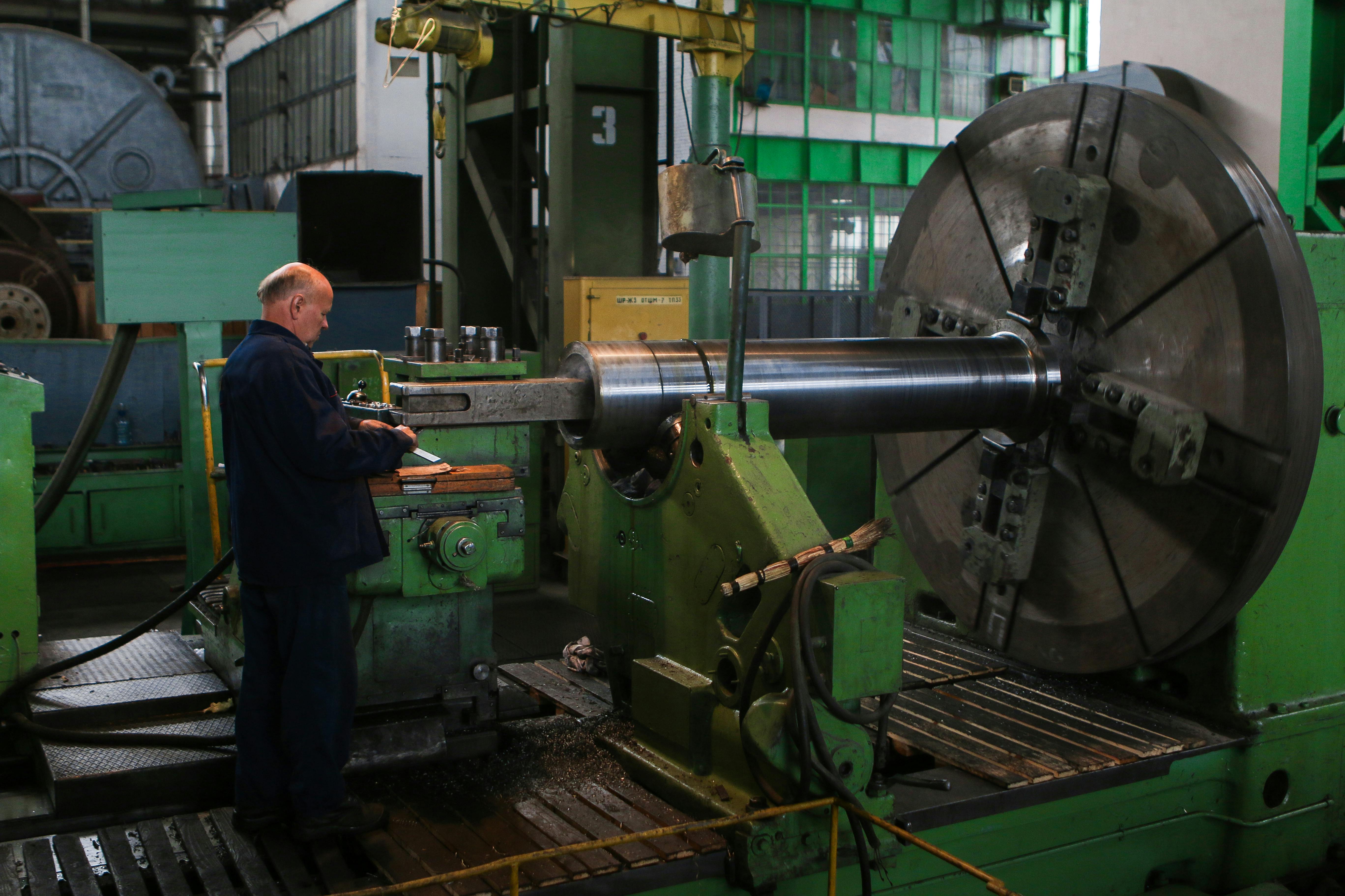 Machinist operating a large industrial lathe to shape a metal cylinder inside a manufacturing facility.