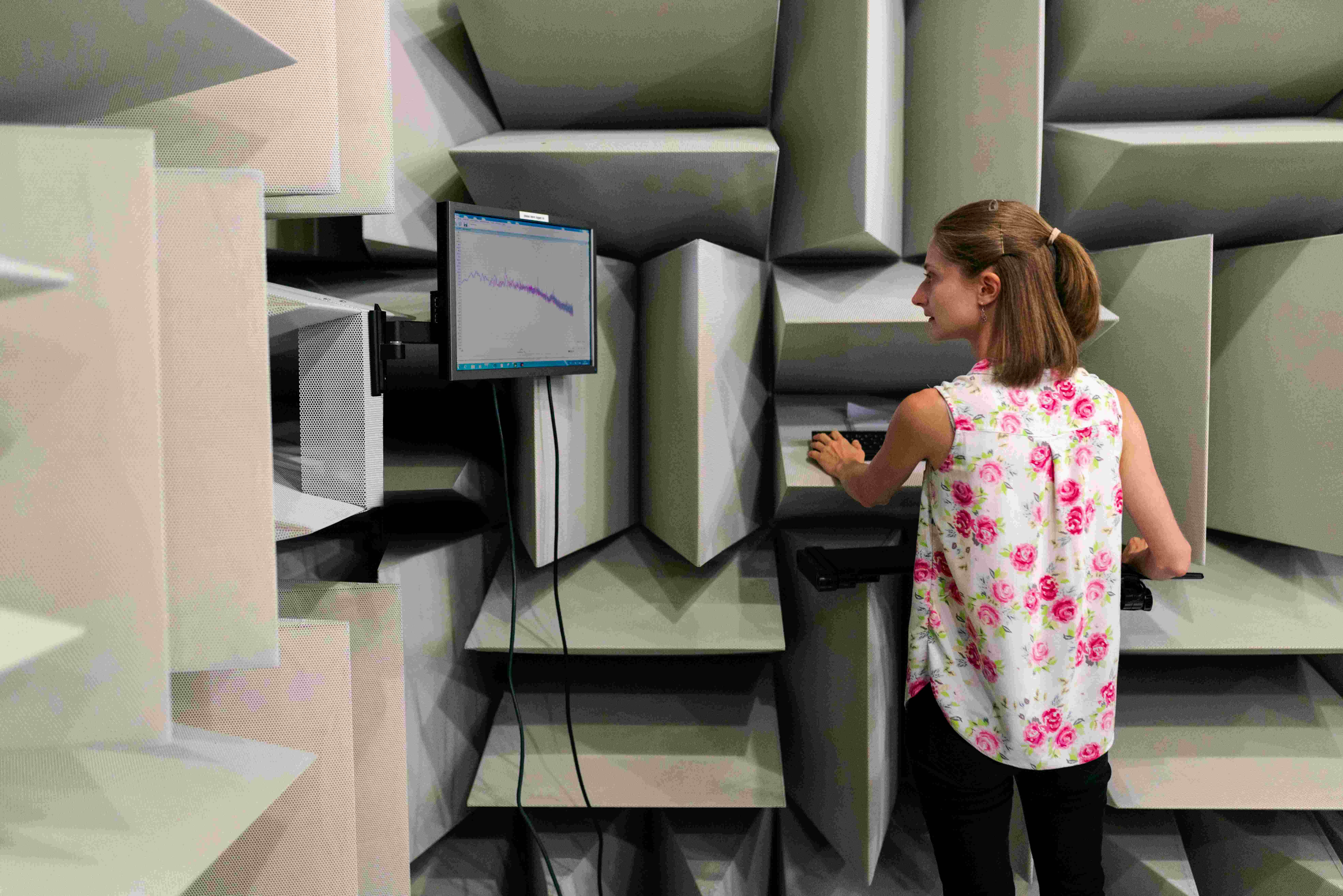 Woman in a sleeveless floral blouse operating a keyboard in an anechoic chamber with a monitor displaying a graph.