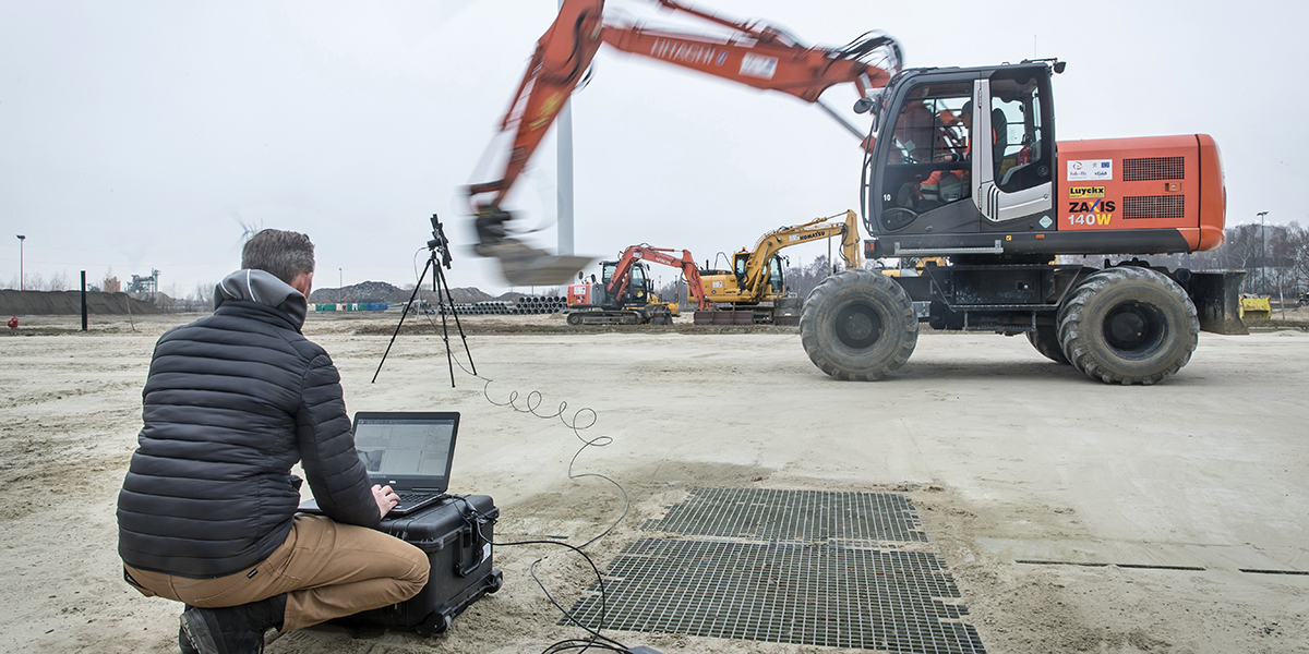 Technician operating a laptop connected to monitoring equipment while observing an excavator in motion at a construction site.