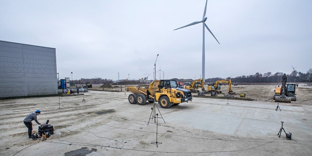Wide view of a construction site with heavy machinery, wind turbines, and technicians operating monitoring equipment on the ground.