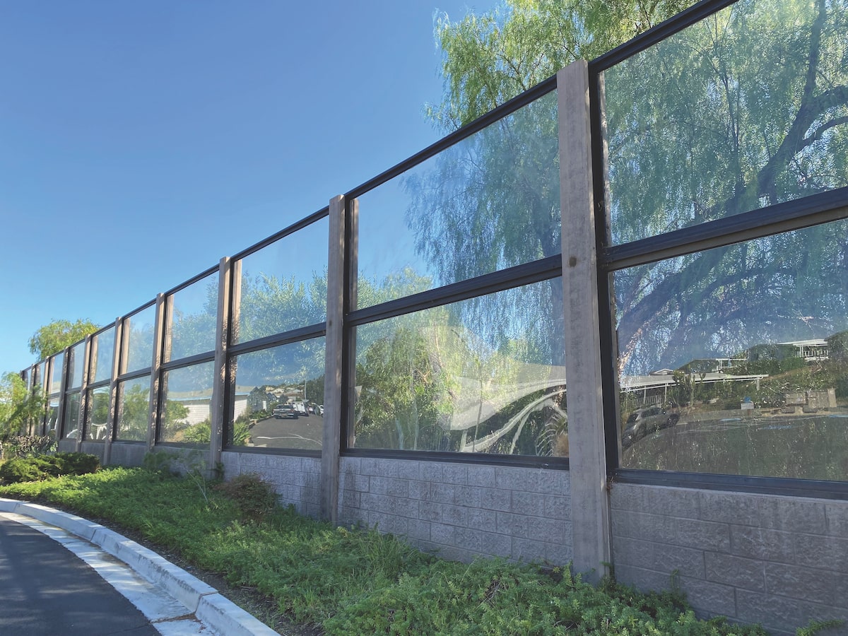 Transparent outdoor acoustic barrier with clear panels mounted on a concrete base, blending into landscaped surroundings for effective noise control.