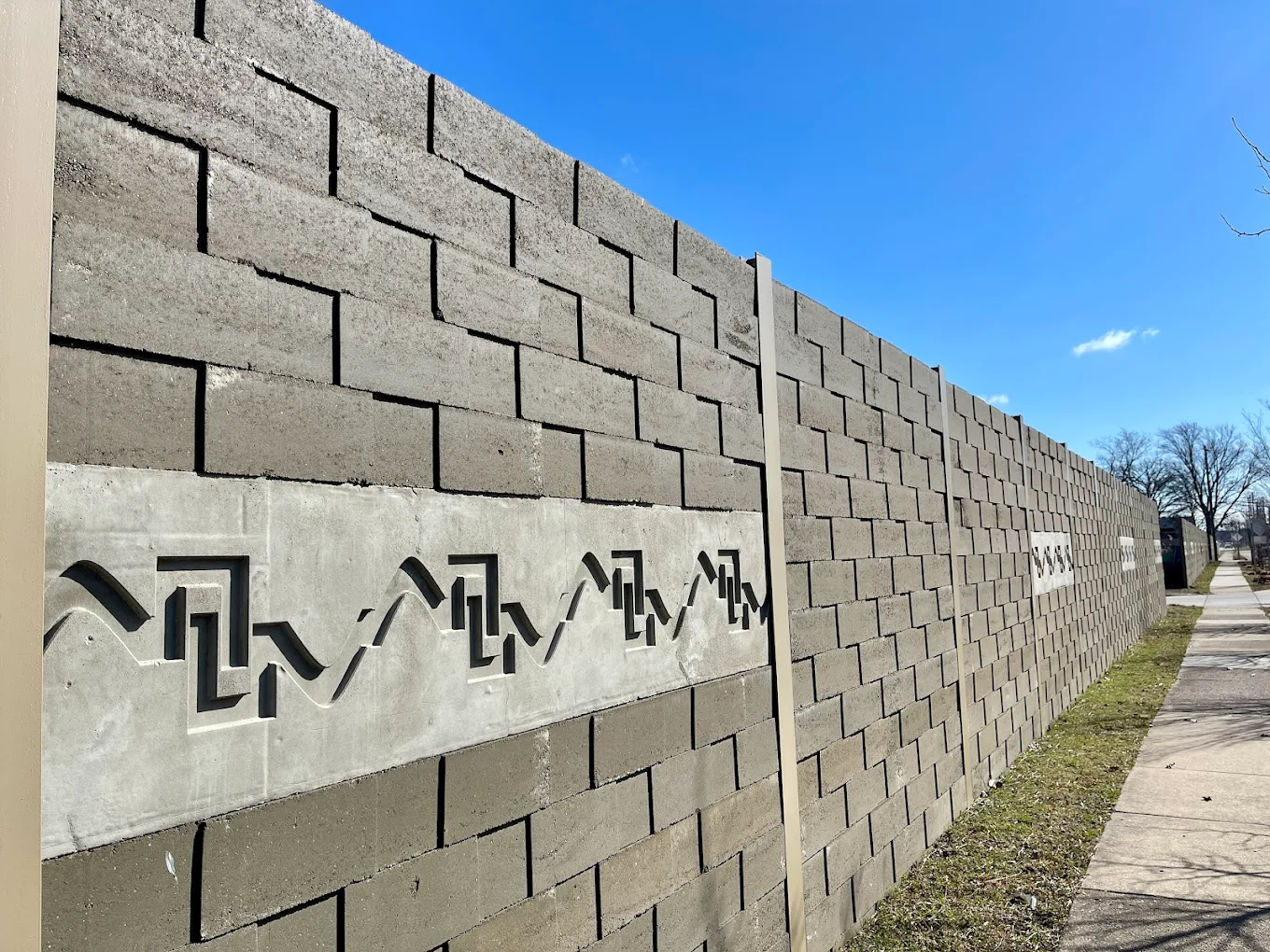 Decorative concrete sound barrier wall along a sidewalk, featuring patterned inlays for visual appeal and effective urban noise control.
