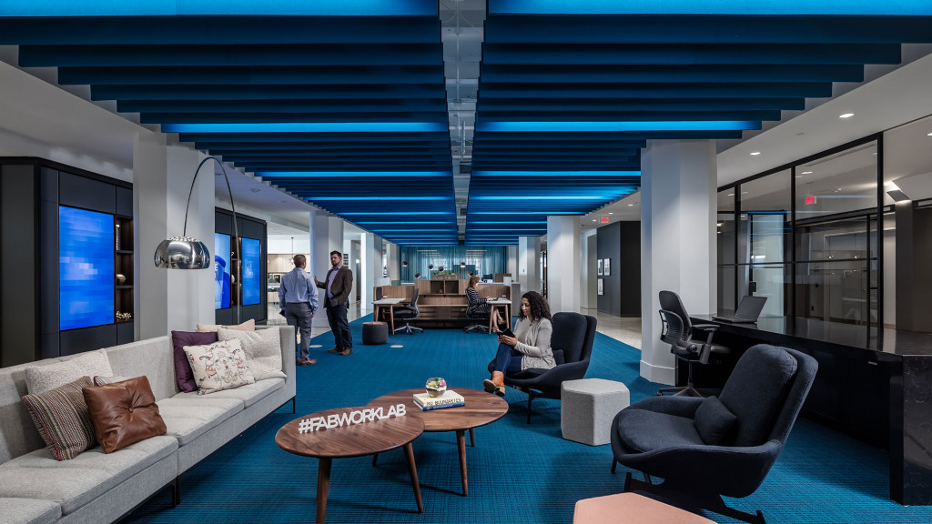 Modern open office lounge with blue acoustic ceiling baffles and collaborative seating areas, showcasing workplace noise control solutions.