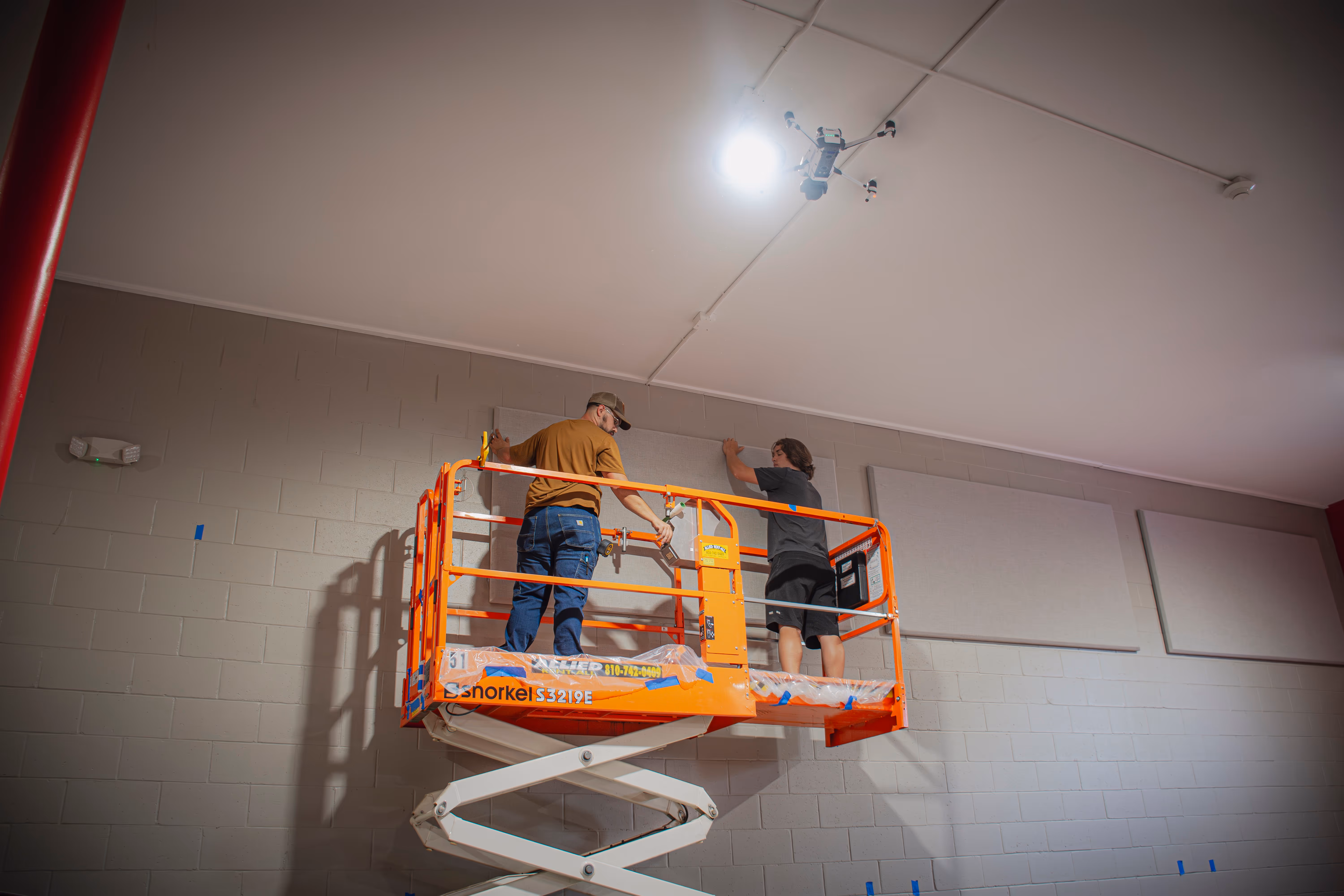 Two men on an orange scissor lift installing panels on a high gray brick wall under bright lighting.