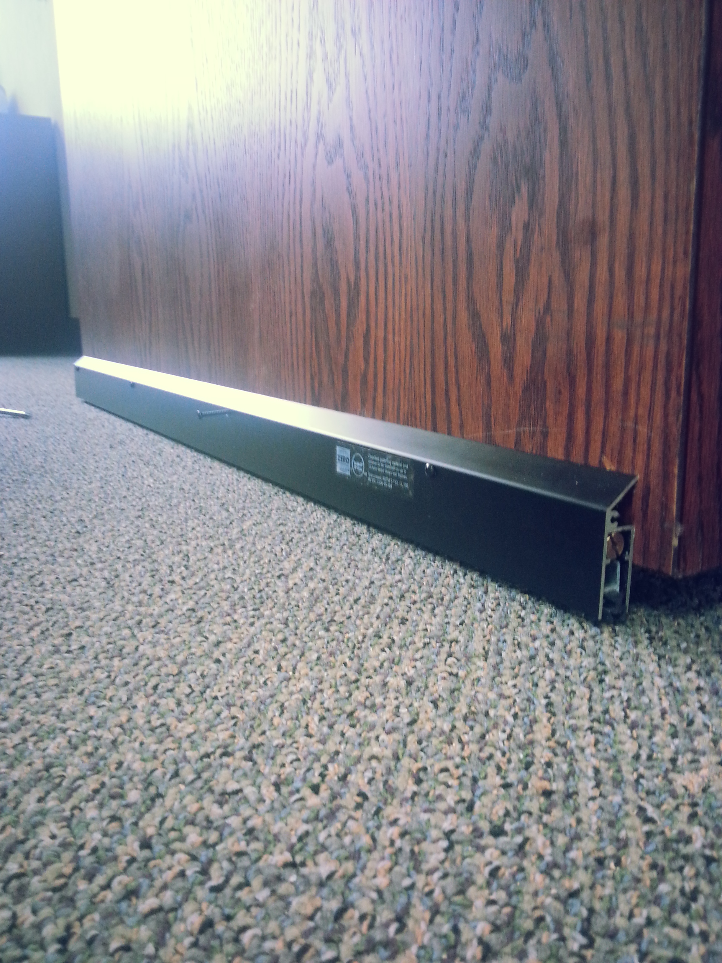 Black metal door bottom seal partially attached to a wooden door on a textured carpet floor.