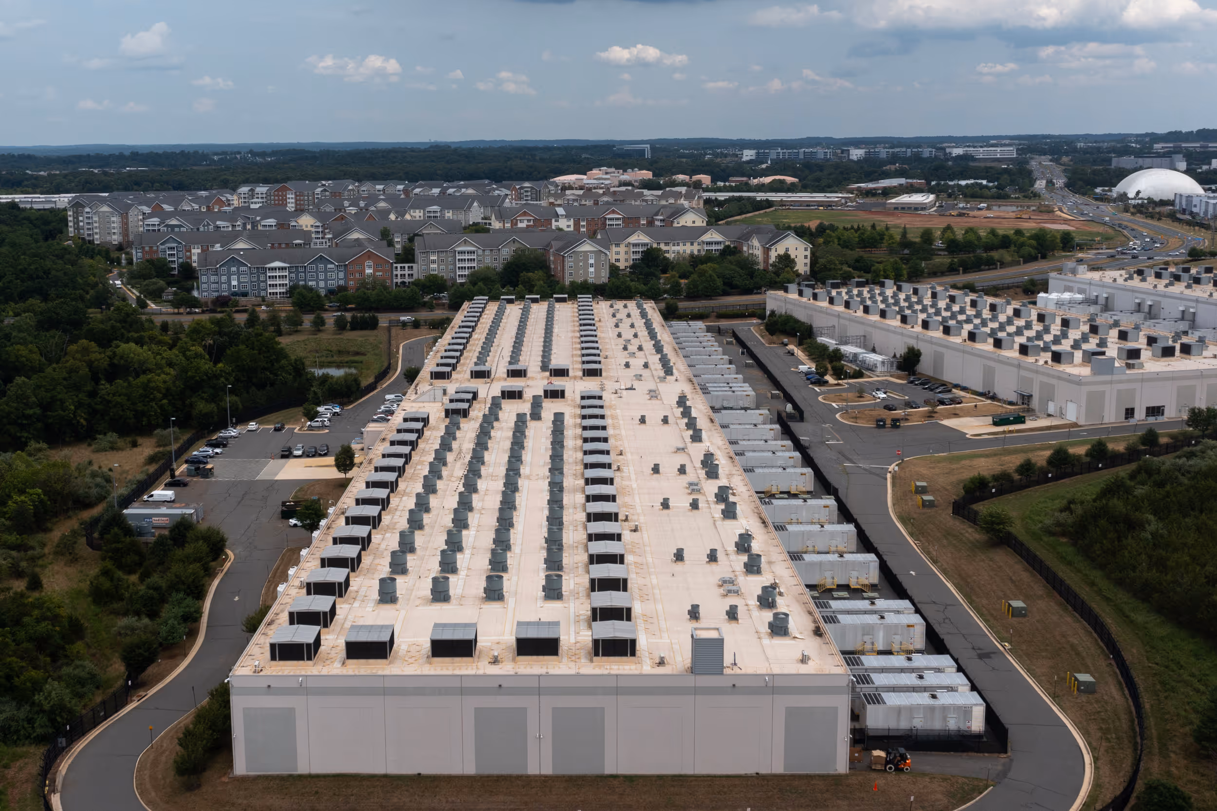 Aerial view of a large industrial building with multiple rooftop ventilators and cooling units, surrounded by roads, parking areas, and residential buildings in the background.