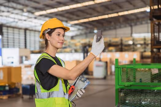 Woman in a yellow hard hat and reflective vest using a handheld device in a warehouse.