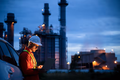 Engineer in safety gear using a laptop outside an industrial power plant at dusk.