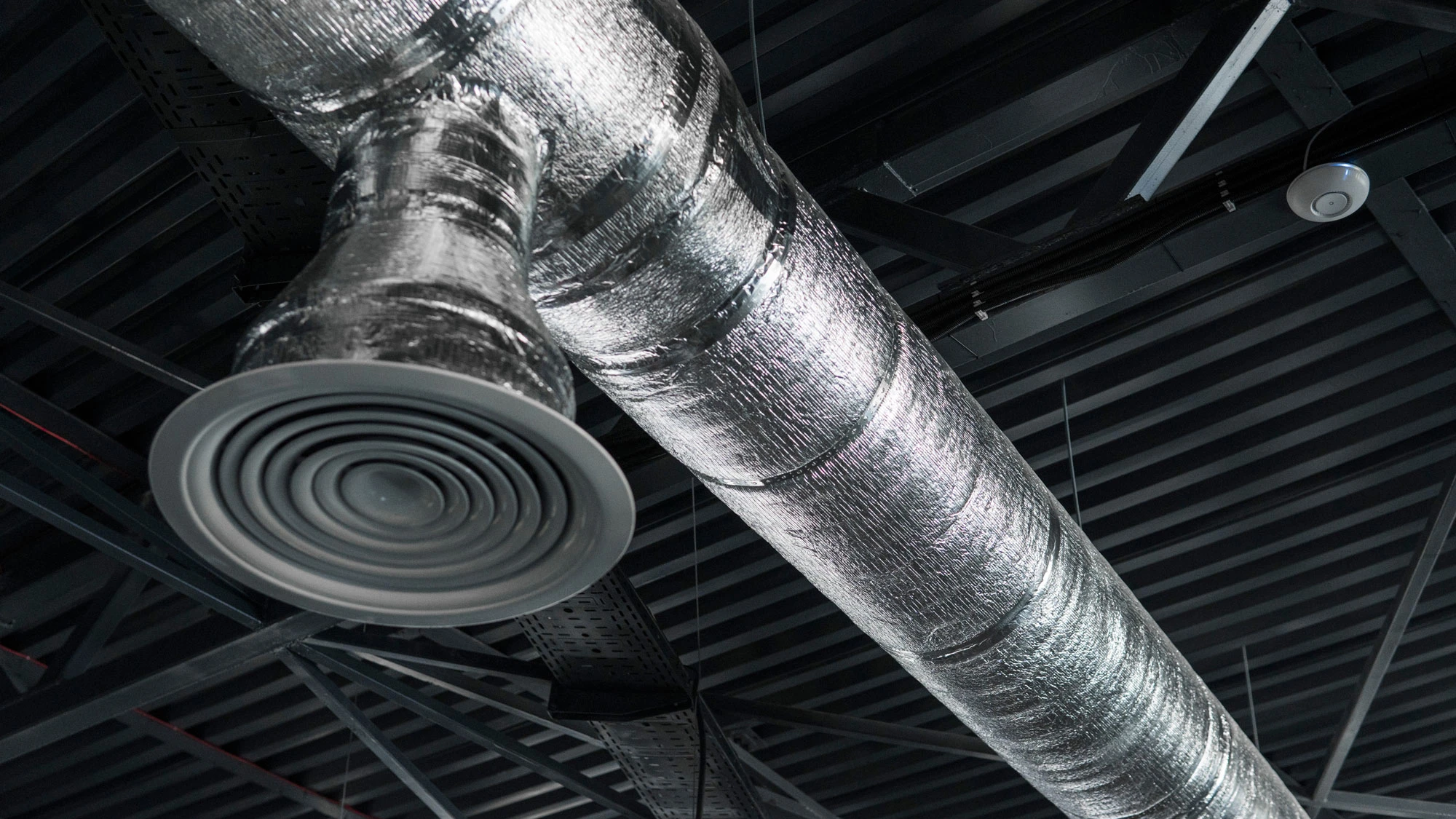 Ceiling vent duct wrapped in acoustical duct wrap in an industrial-style ceiling with metal beams.