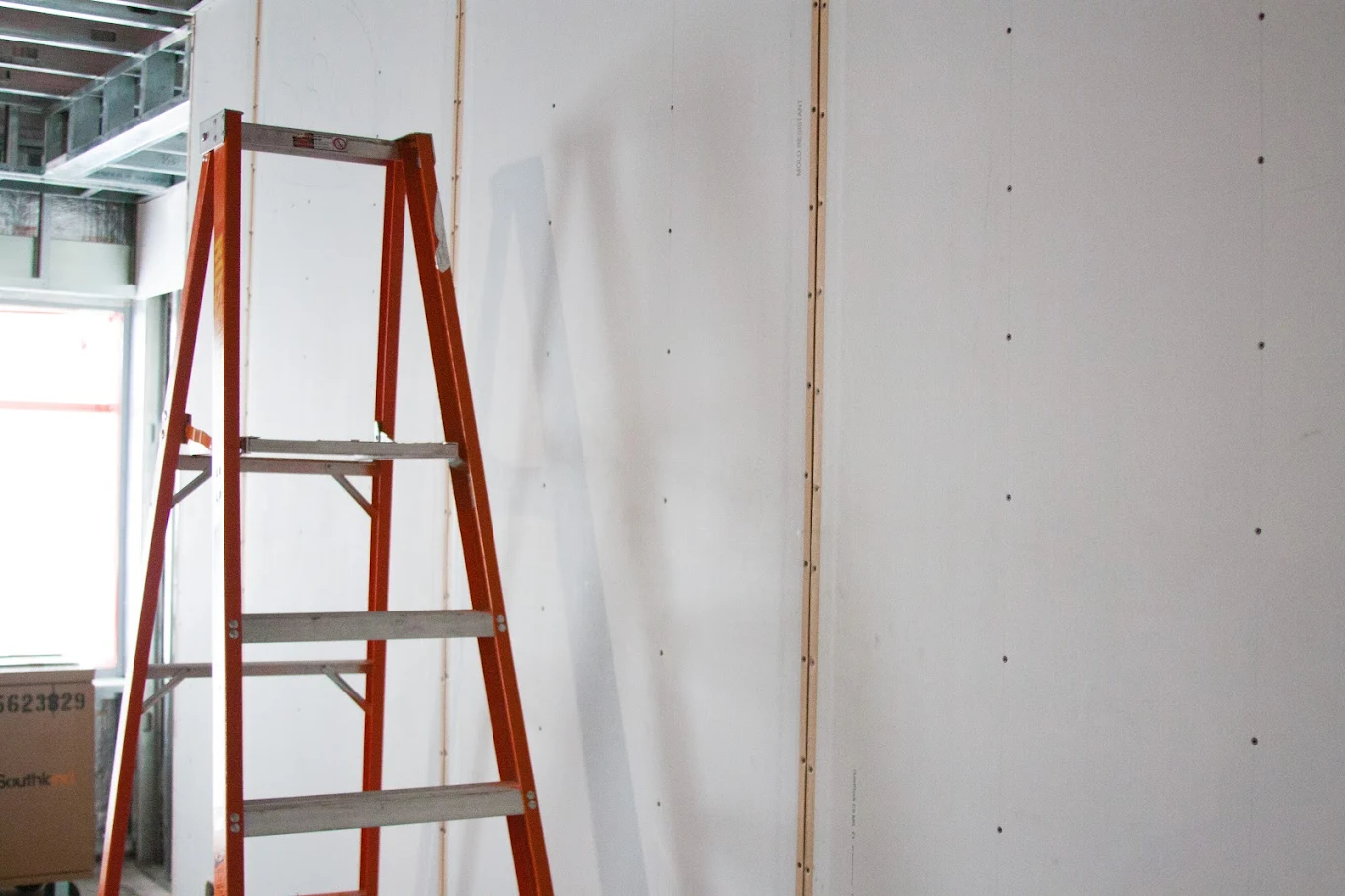 Orange step ladder leaning against an unfinished white drywall interior wall in a construction site.