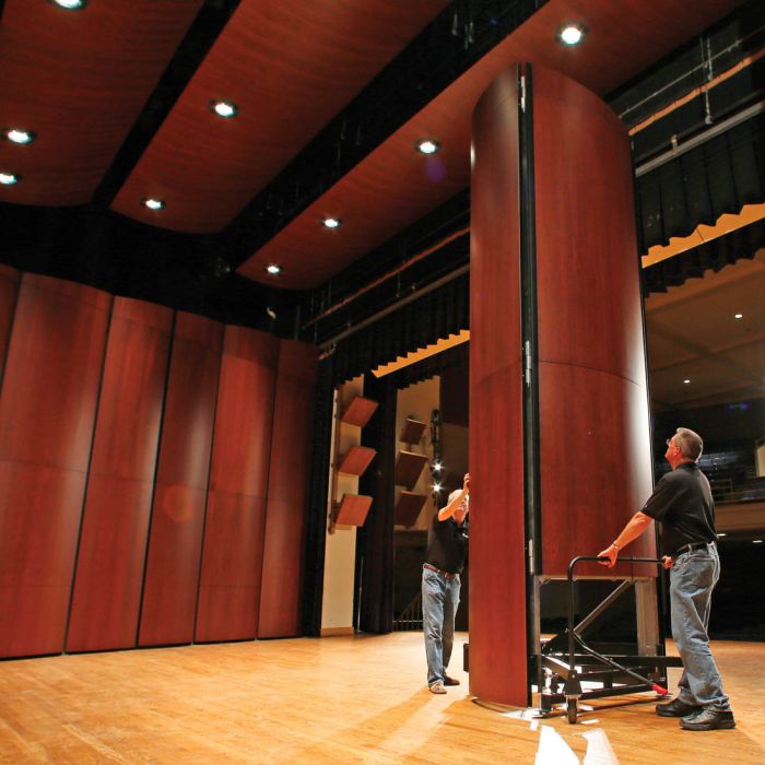 Two men adjusting a large curved wooden acoustic shell on a stage inside a theater.