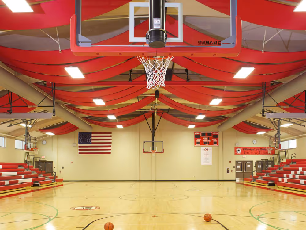 Empty indoor basketball court with red and white bleachers, hanging basketball hoops, and an American flag on the wall.