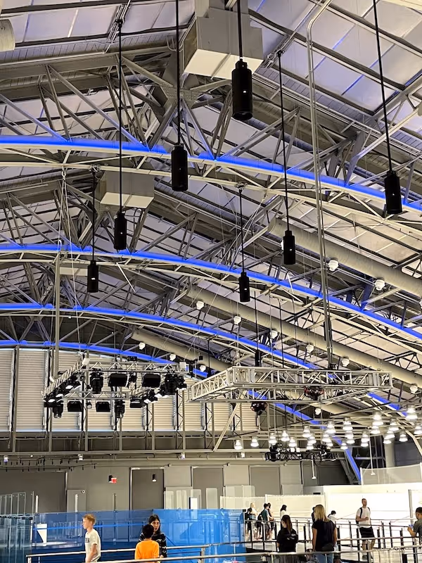 Ceiling view of an indoor sports facility with hanging lights, blue accent lighting, and people walking below.