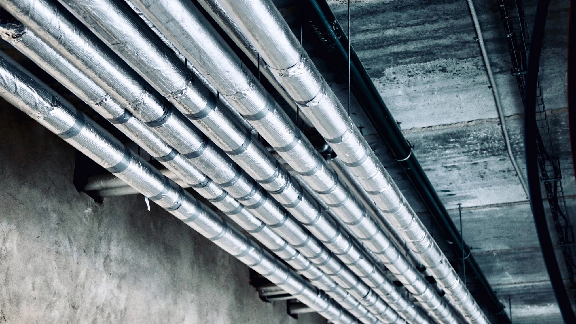 Rows of shiny metal pipes running parallel along a concrete ceiling in an industrial setting.